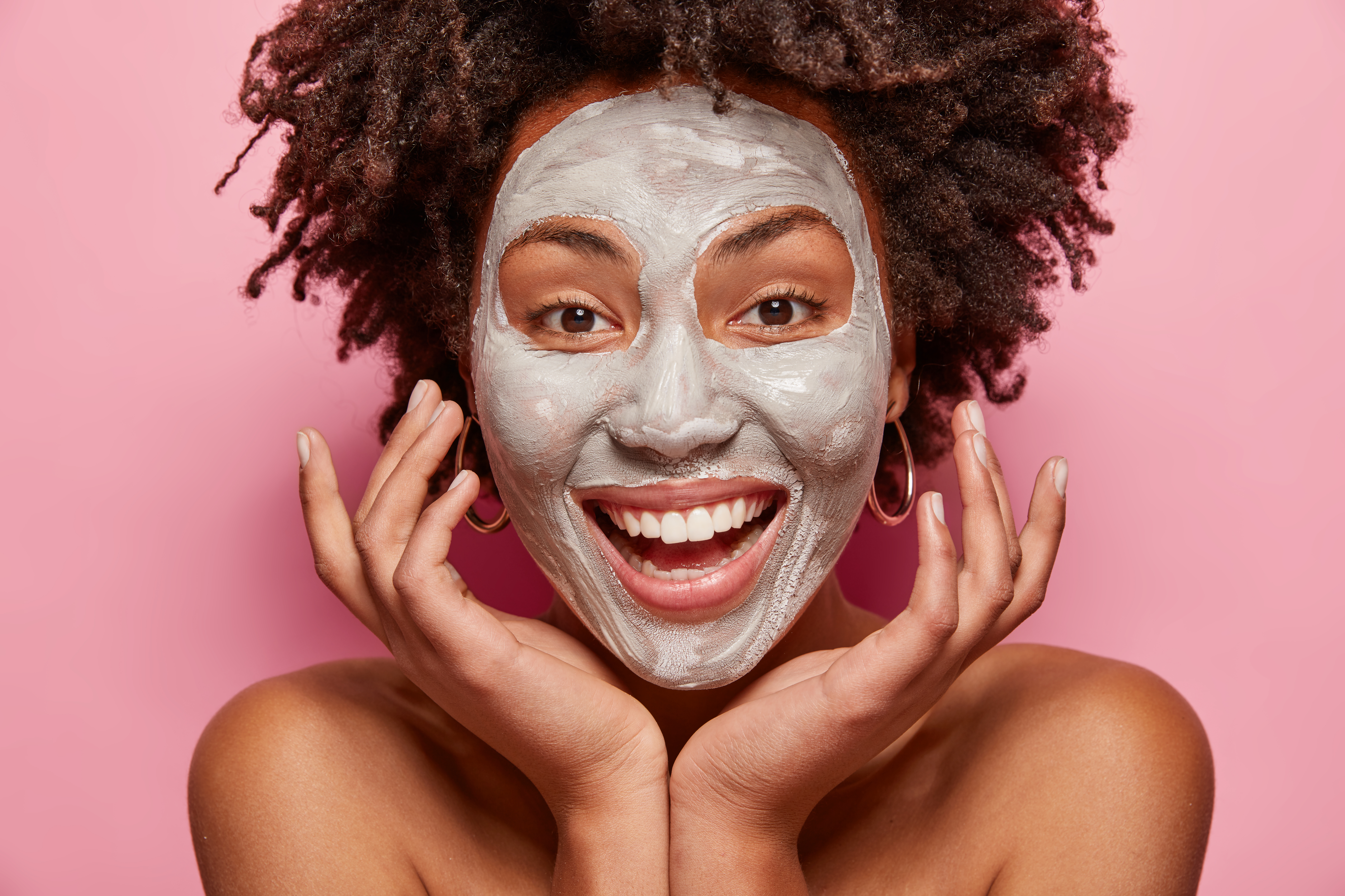 Headshot of cheerful girl with white clay mask, touches face, takes care of skin and beauty, has positive smile, Afro haircut, models over pink background, poses indoor. Facial treatment concept